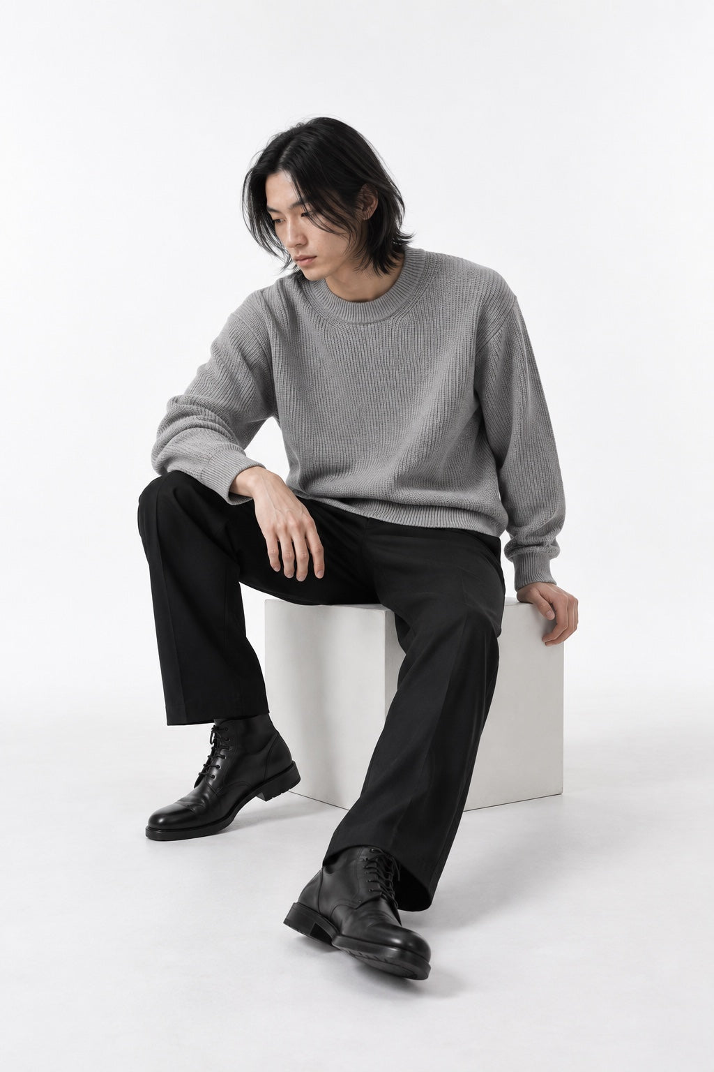 Full-body long-haired male model seated in the light-grey Trace Crewneck Knit in a white studio