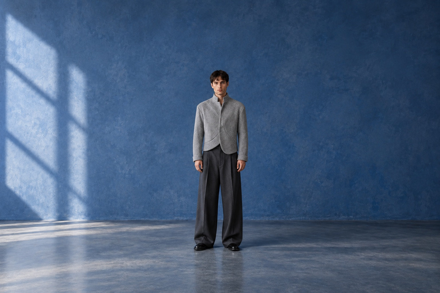 Full-body male model wearing the gray Soren Asymmetric Cropped Jacket in a sunlit blue studio